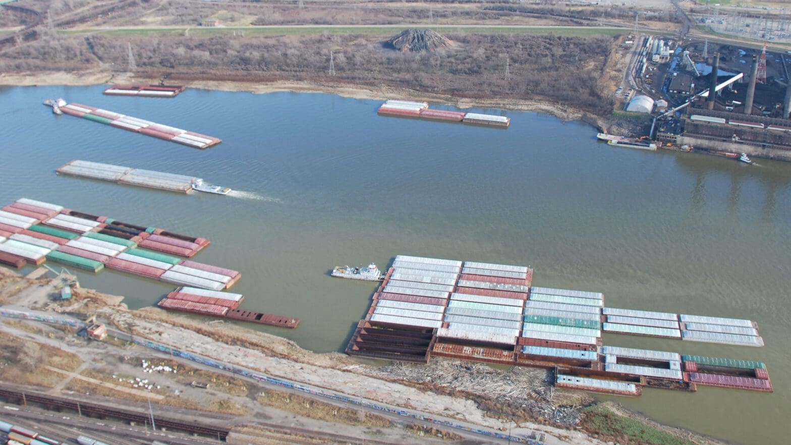 A tug pushing river barges on a river with an industrial plant on the shore.