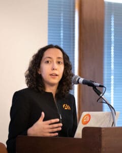 Female speaker in black jacket addressing audience with microphone.