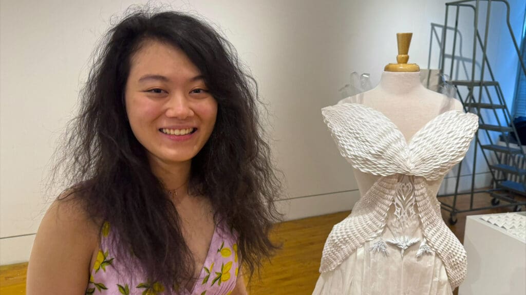 A woman, in the foreground, stands next to a wedding dress in the background